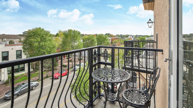 A balcony with a wrought iron railing, a round table with chairs, and a view of a street with cars and buildings.