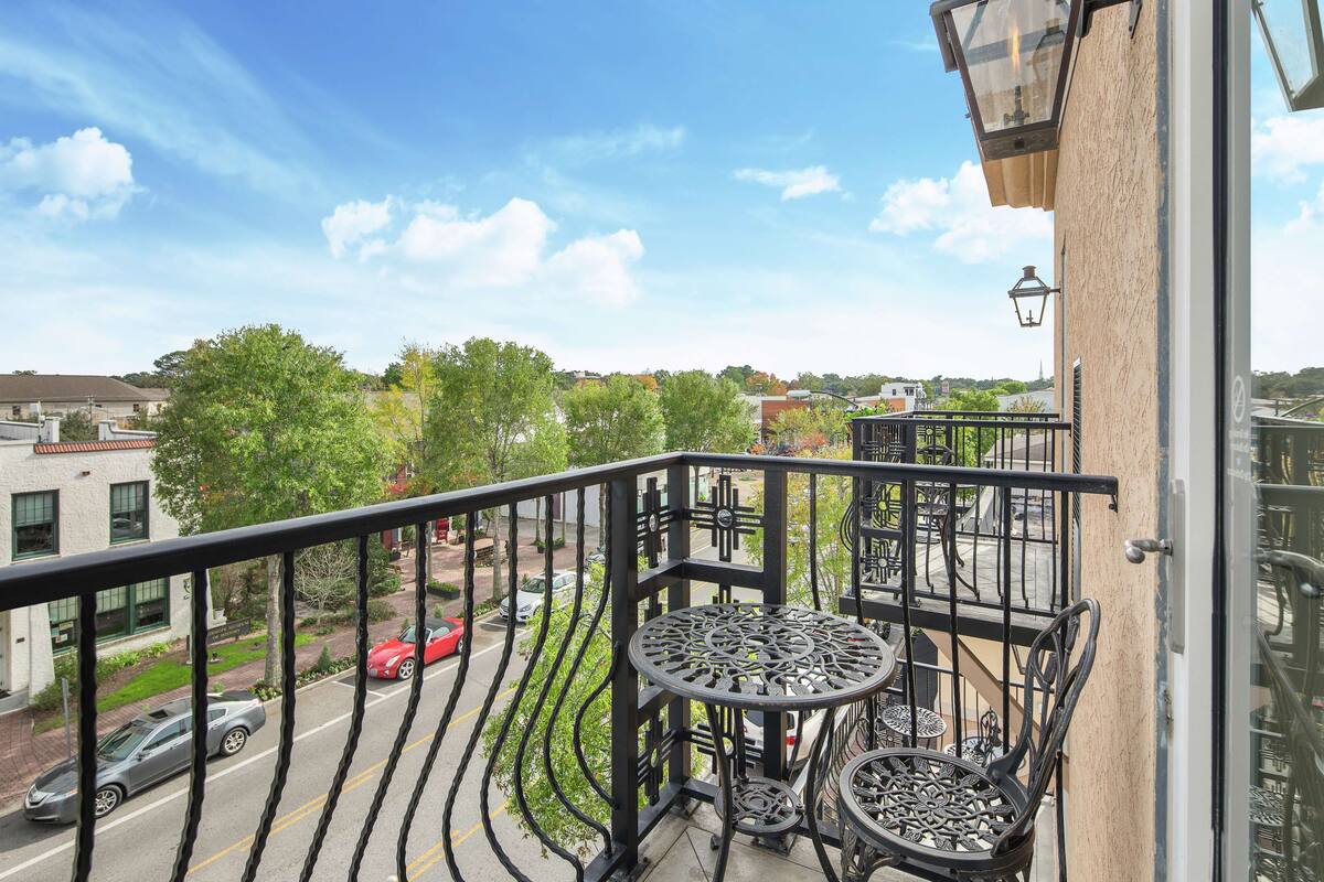 A balcony with a wrought iron railing, a round table with chairs, and a view of a street with cars and buildings.