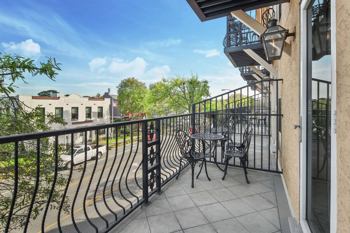 A balcony with a metal railing, outdoor seating, and a view of the street and trees.