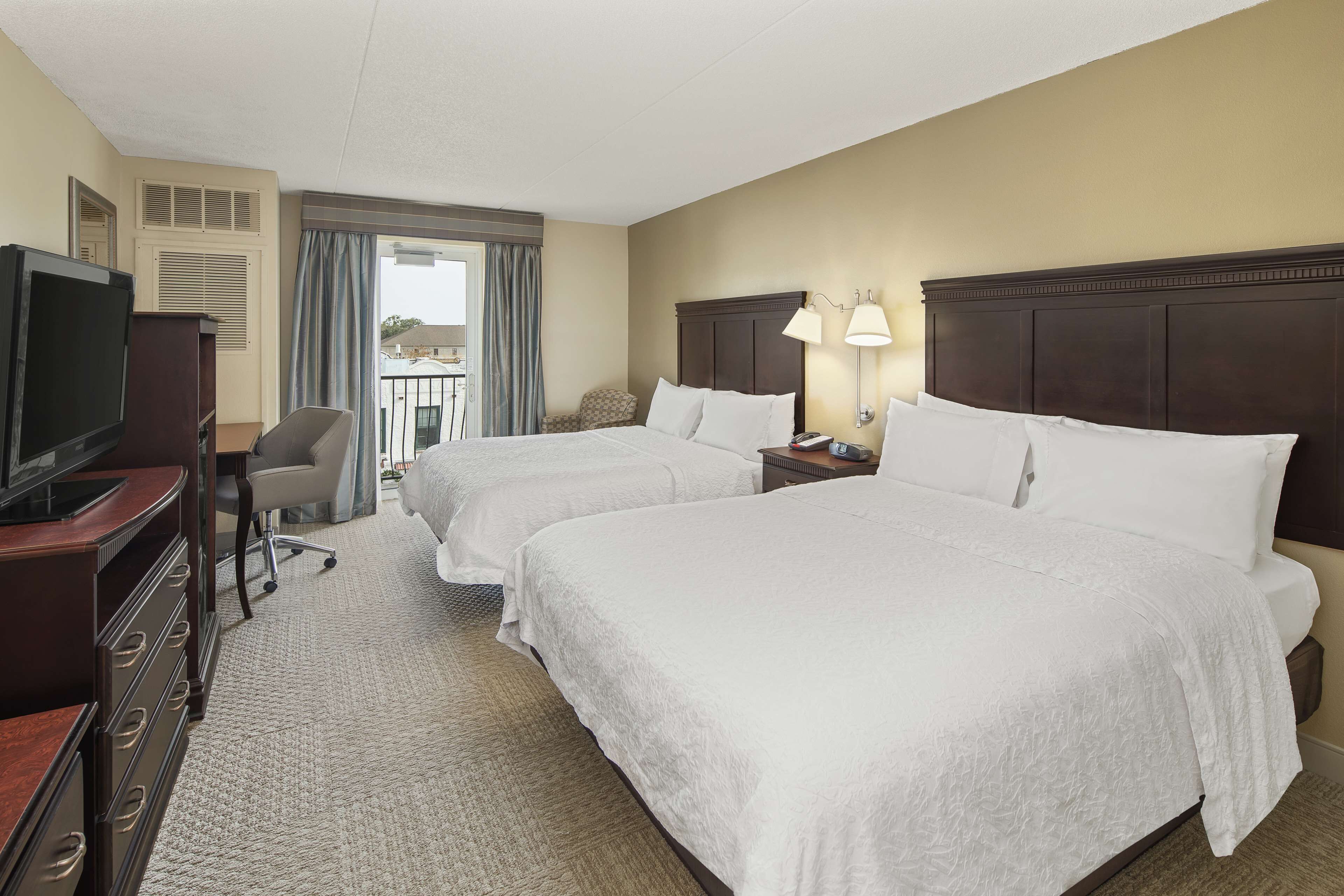 A hotel room with two beds, a desk with a television, a chair, and a balcony with curtains.