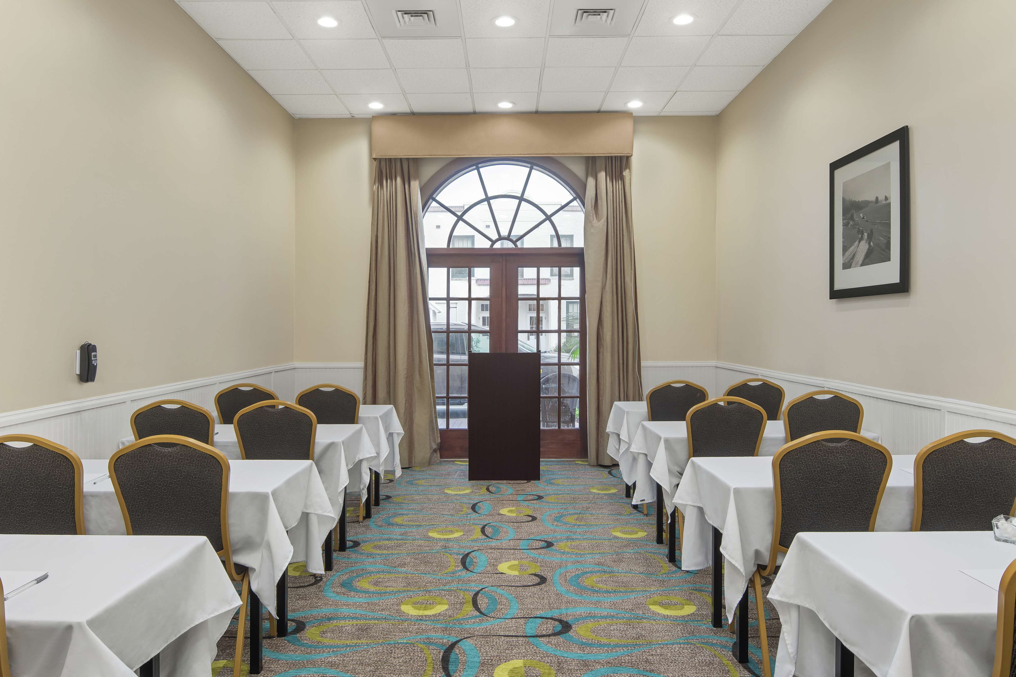 A conference room with rows of tables and chairs, a large window with curtains, and a framed picture on the wall.
