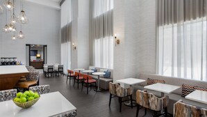 Eine moderne Hotelhalle mit verschiedenen Sitzgelegenheiten, darunter eine Bar, ein Sitzbereich mit Tischen und Stühlen sowie ein Buffet mit einer Obstschale.