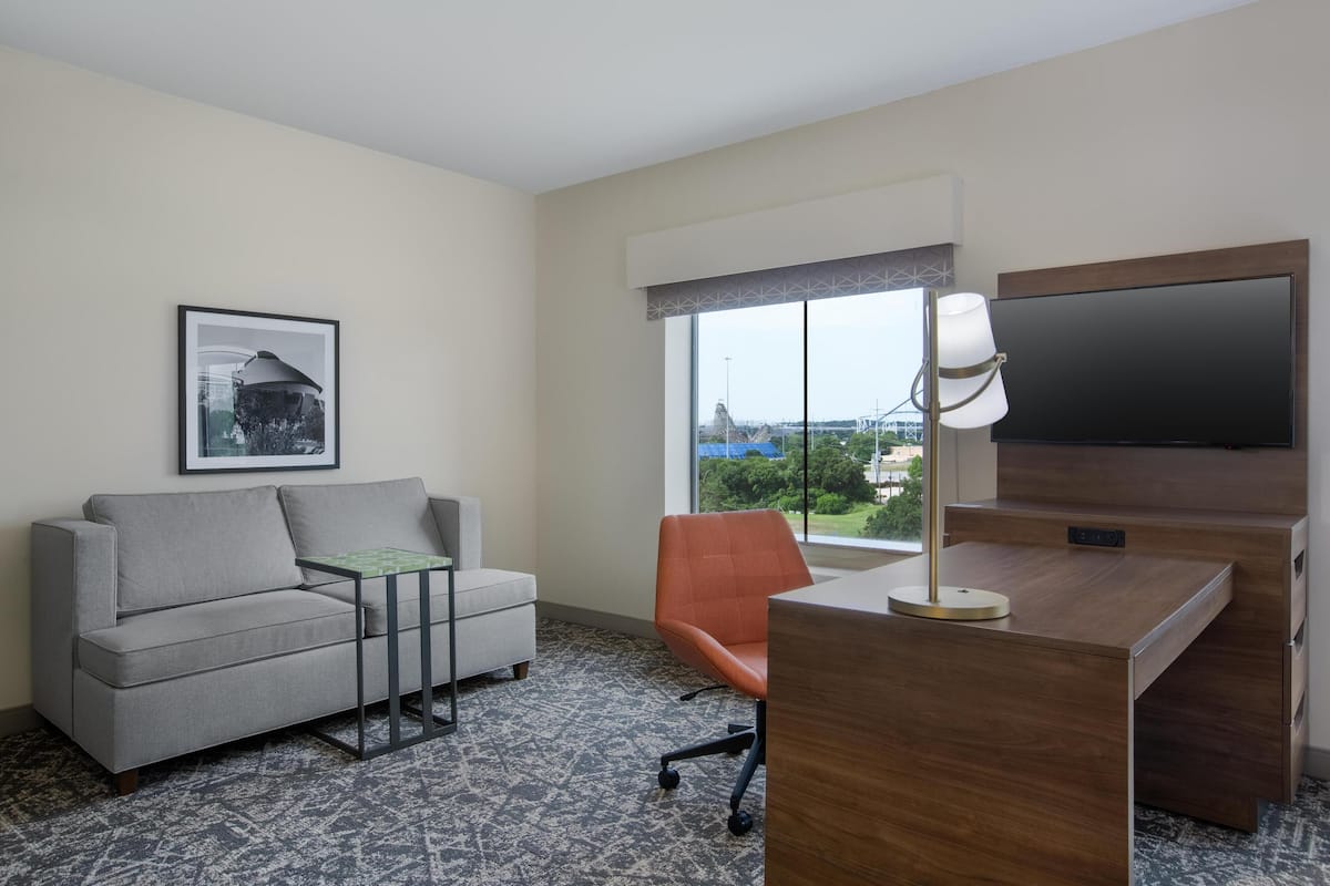 Une chambre d’hôtel avec un canapé gris, une petite table, une chaise orange, un bureau avec une télévision et une grande fenêtre donnant sur la ville.