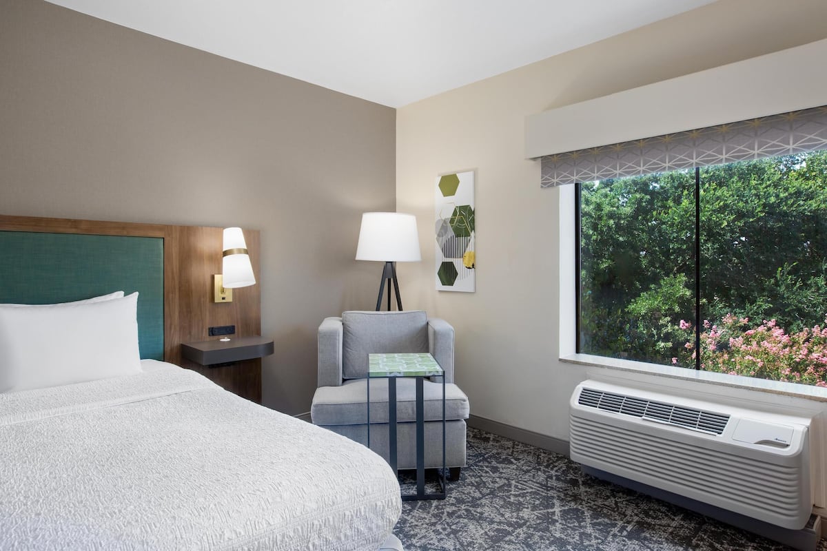 Une chambre d’hôtel équipée d’un lit, d’une chaise, d’une table, d’une lampe, d’une fenêtre donnant sur un espace verdoyant et d’un climatiseur fixé au mur.