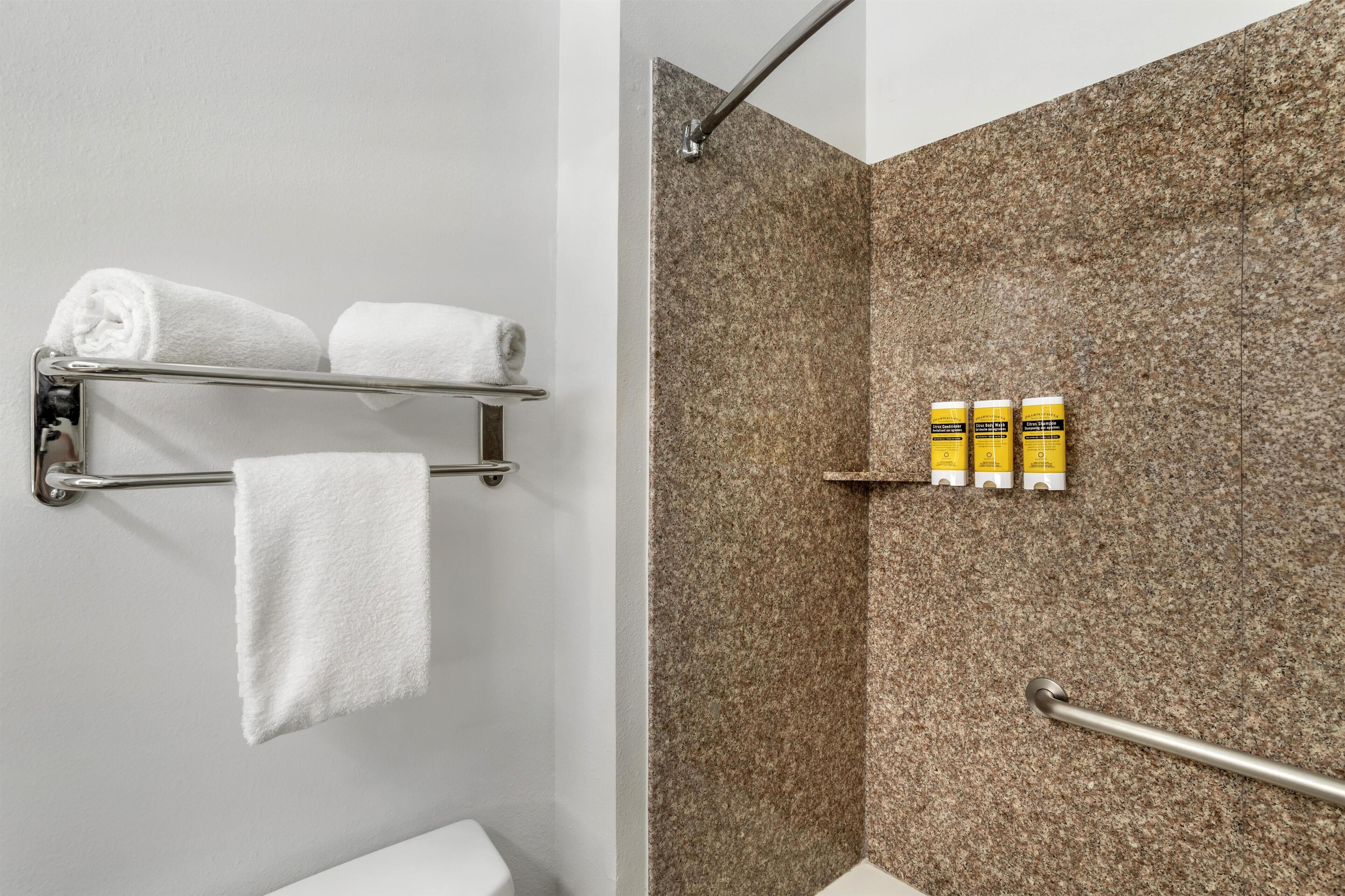 A bathroom with a shower area, a towel rack with two towels, and three bottles on a shelf.