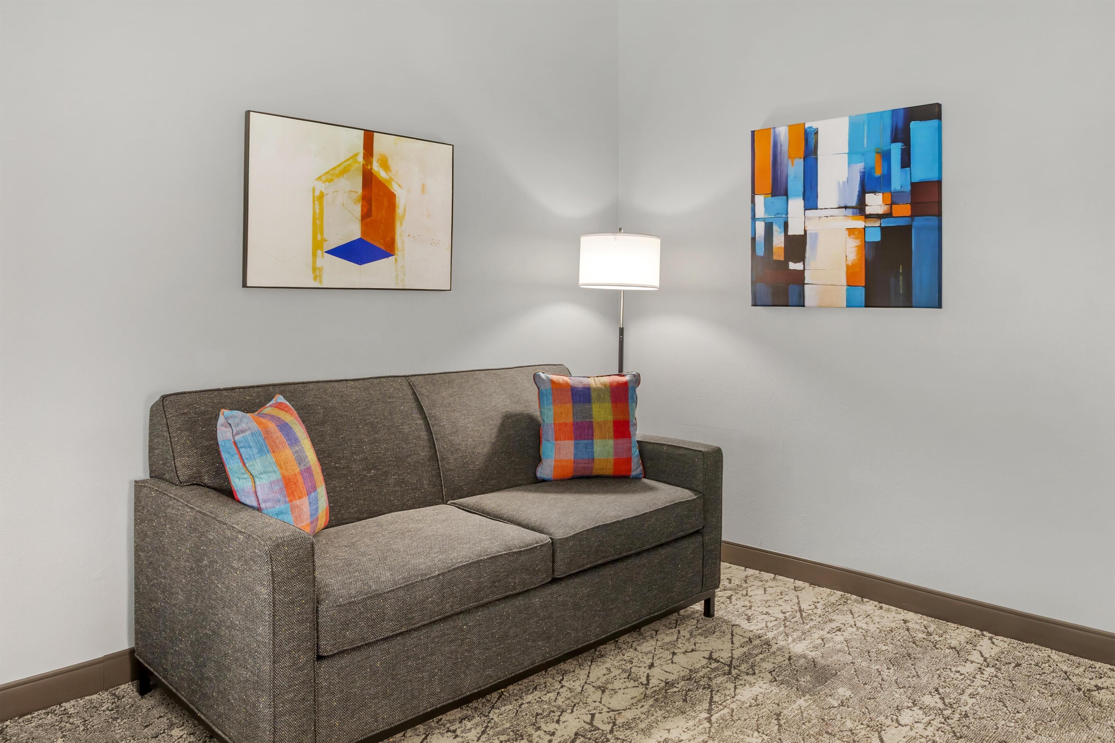Un canapé gris agrémenté de deux coussins colorés, un lampadaire et deux tableaux abstraits accrochés au mur.
