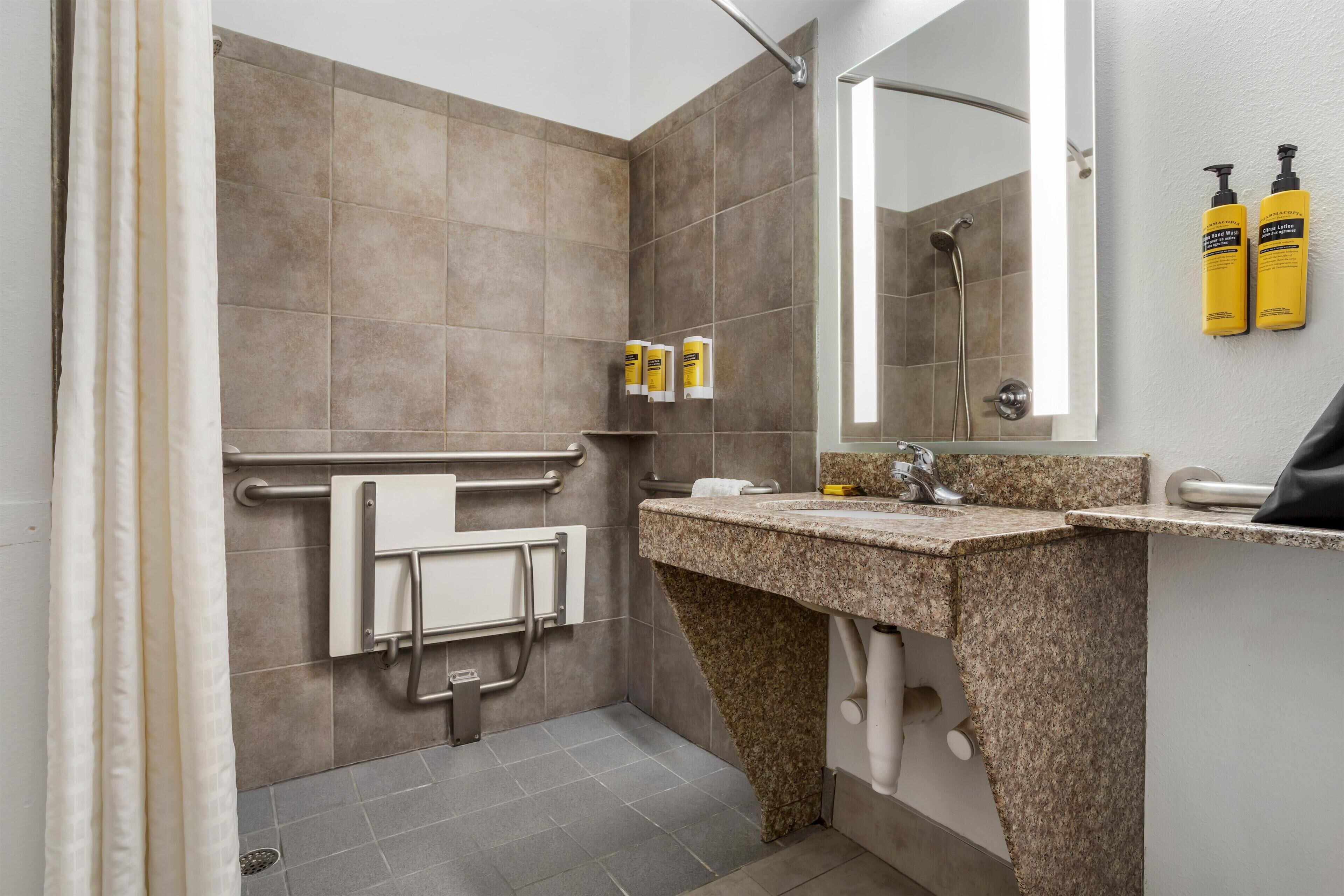 A modern bathroom with a shower, grab bars, a sink with a mirror, and two yellow bottles on the wall.