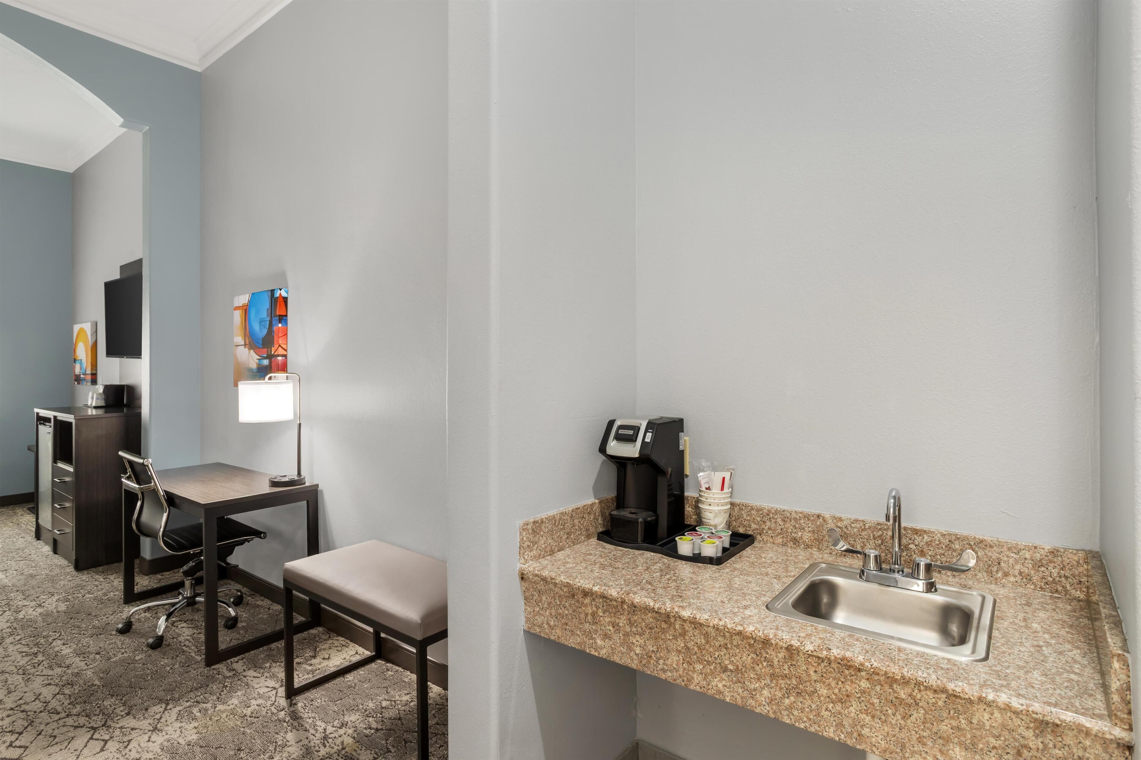 A hotel room with a desk, chair, and a small kitchenette area with a coffee maker and sink.