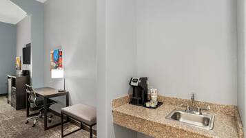 A hotel room with a desk, chair, and a small kitchenette area with a coffee maker and sink.