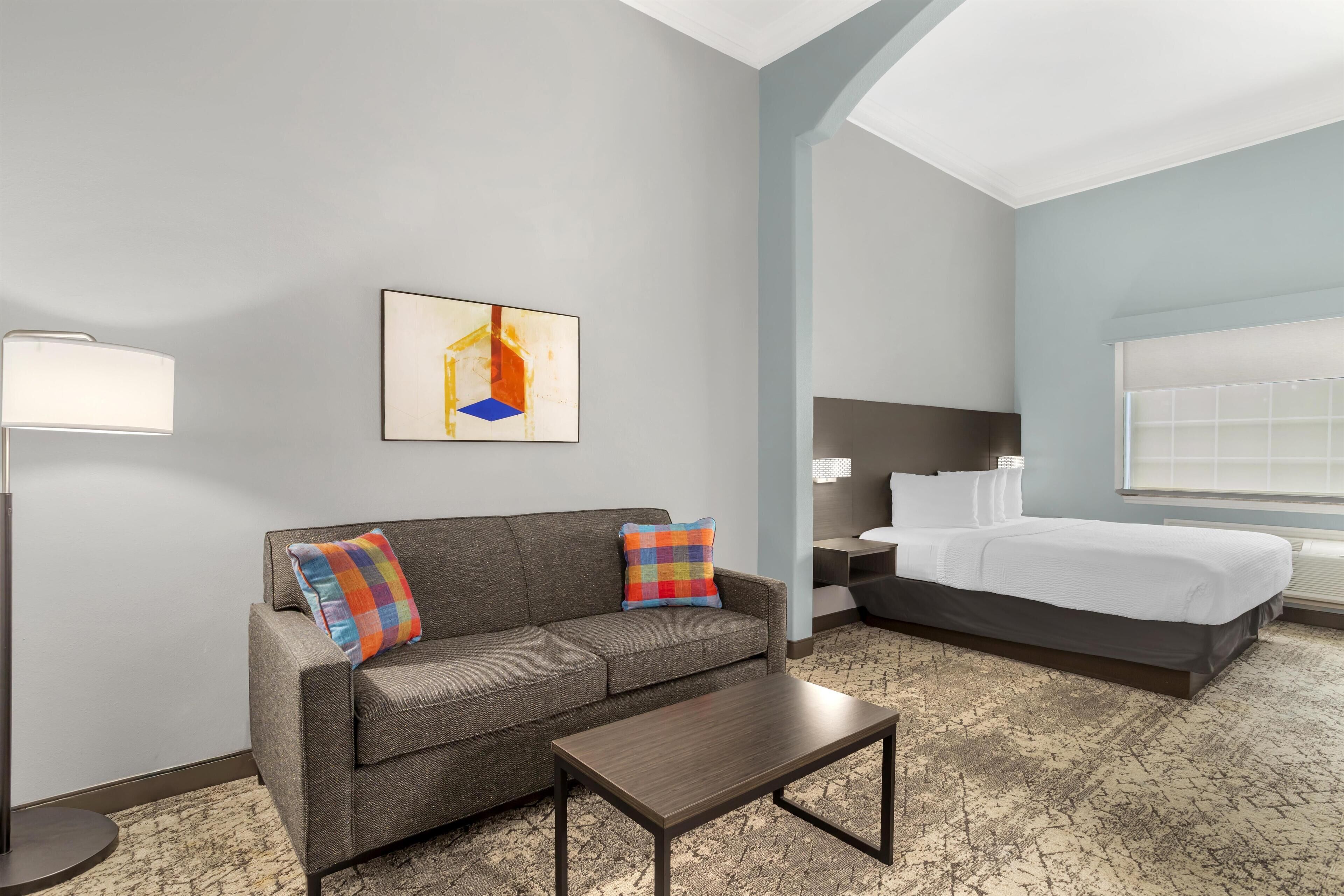 A hotel room with a gray sofa, a small table, a bed with white bedding, and a painting on the wall.