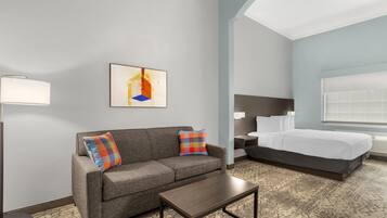 Une chambre d’hôtel avec un canapé gris, une petite table, un lit avec une literie blanche et un tableau au mur.