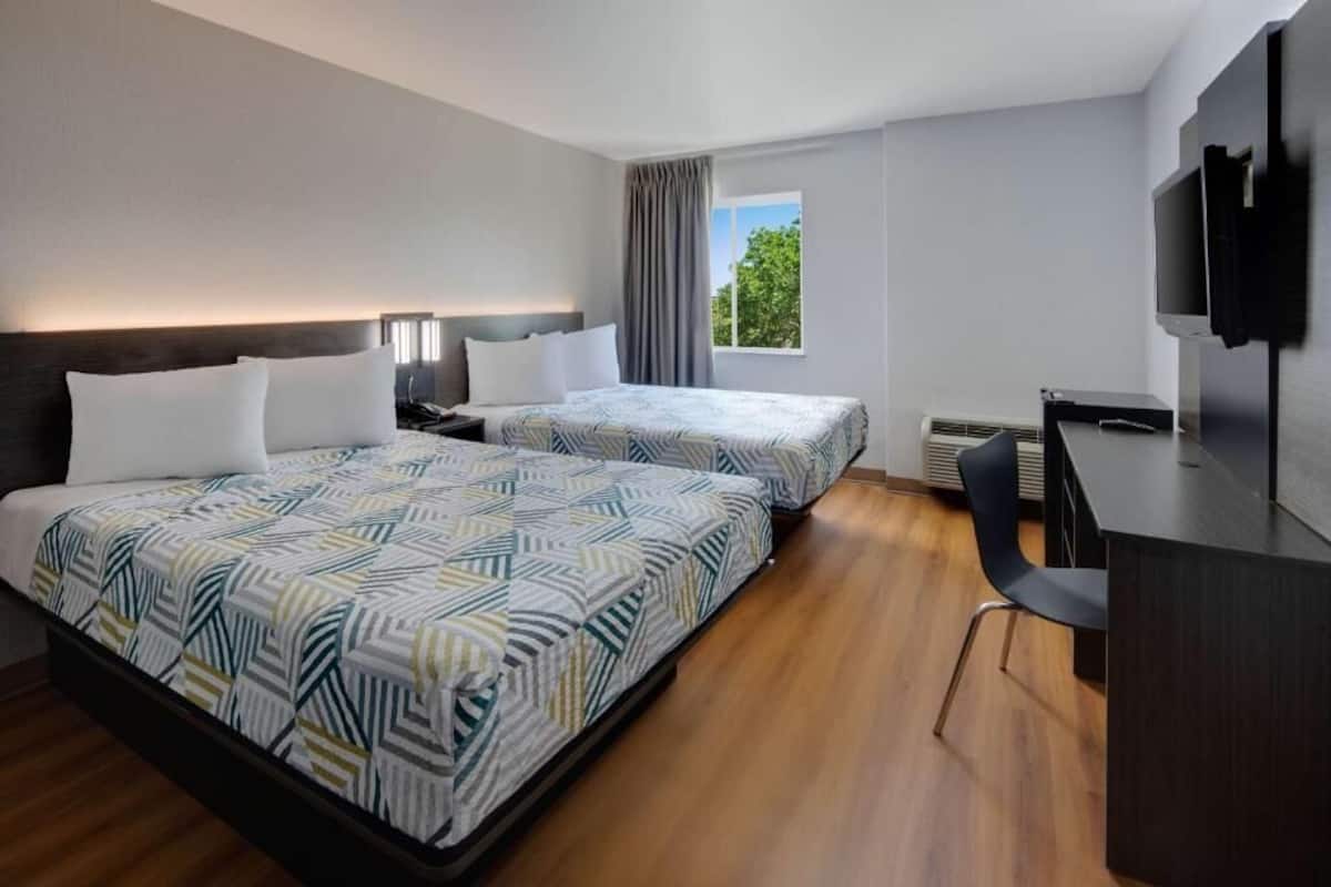Une chambre d’hôtel avec deux lits, un bureau, une chaise, une télévision et une fenêtre avec des rideaux.