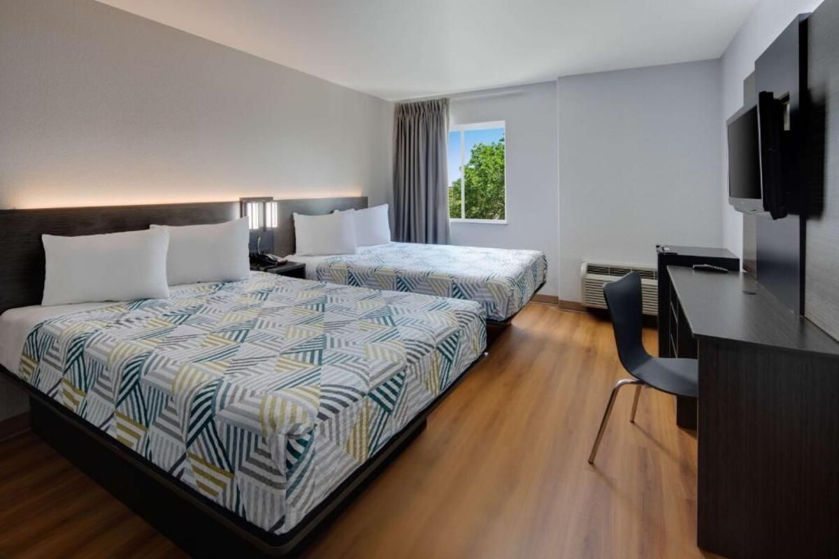 Habitación de hotel con dos camas, un escritorio, una silla, un televisor y una ventana con cortinas.