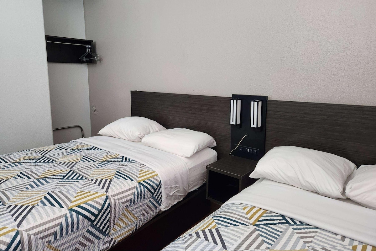Une chambre d’hôtel avec deux lits simples, une table de chevet, une étagère fixée au mur et un miroir.