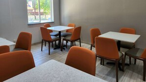 A dining area with tables and chairs, a window with a view of greenery and buildings, and a light-colored floor.