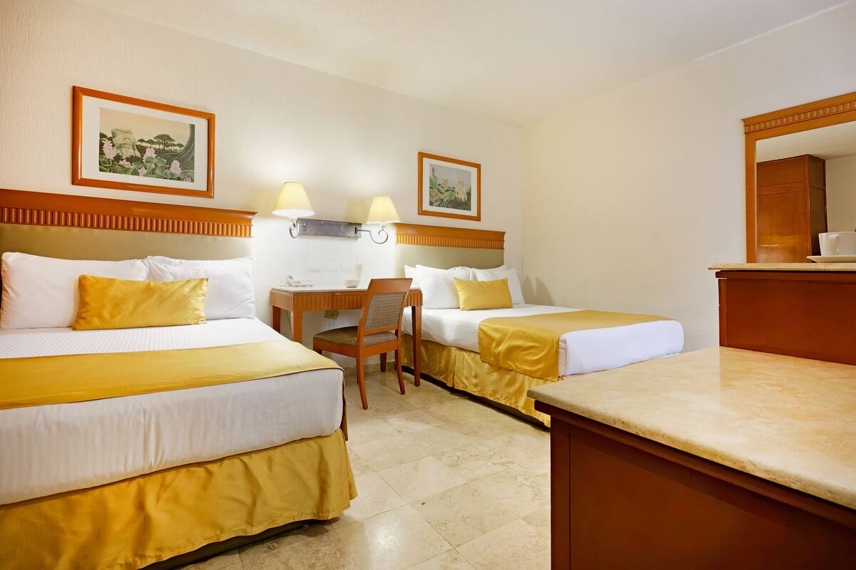 A hotel room with two beds, a desk, and a chair. There are two framed pictures on the wall.