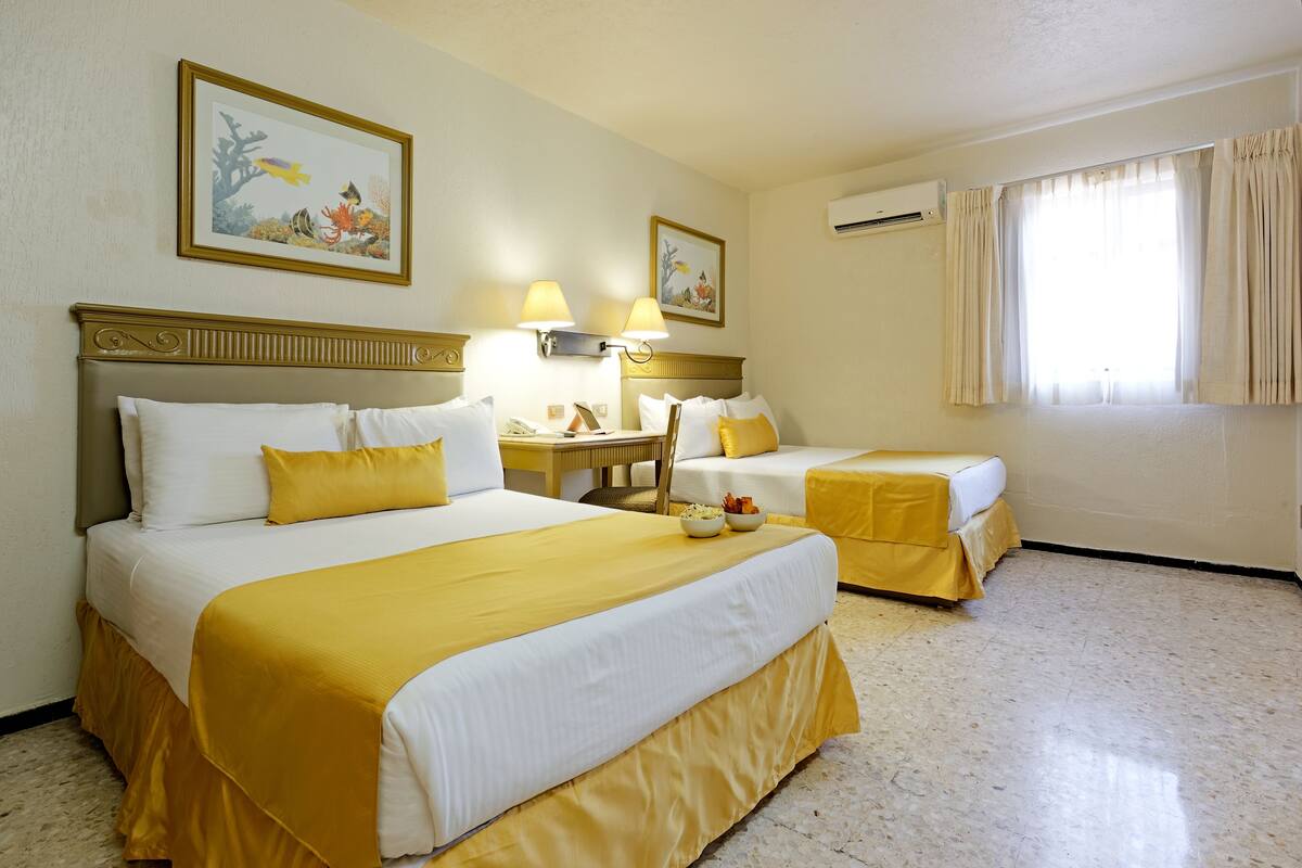 A hotel room with two beds, a desk, and a chair. There are two paintings on the walls and an air conditioning unit on the wall.