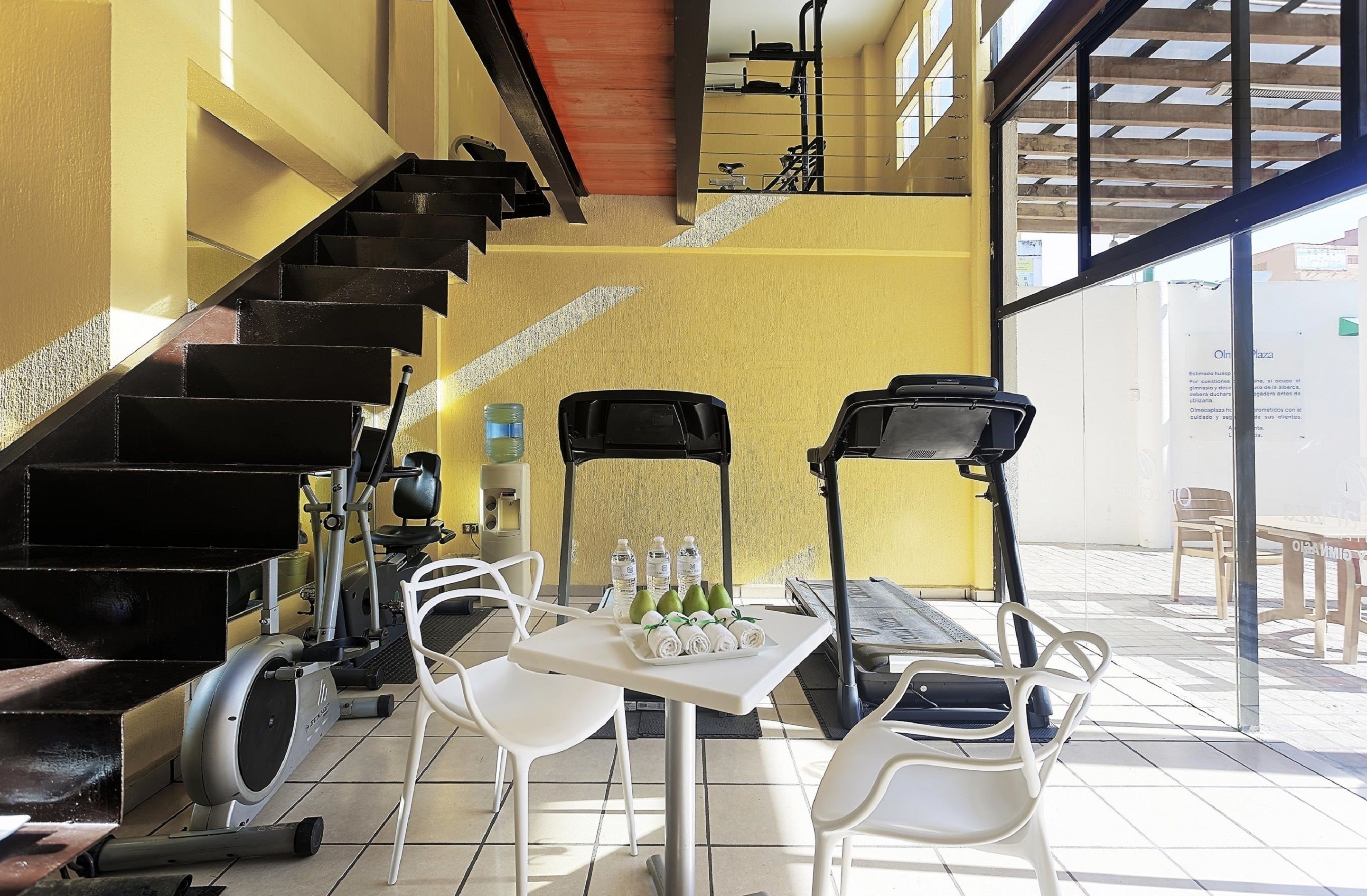 Un área de gimnasio con equipamiento de ejercicio, una mesa con botellas de agua y toallas, y una escalera.