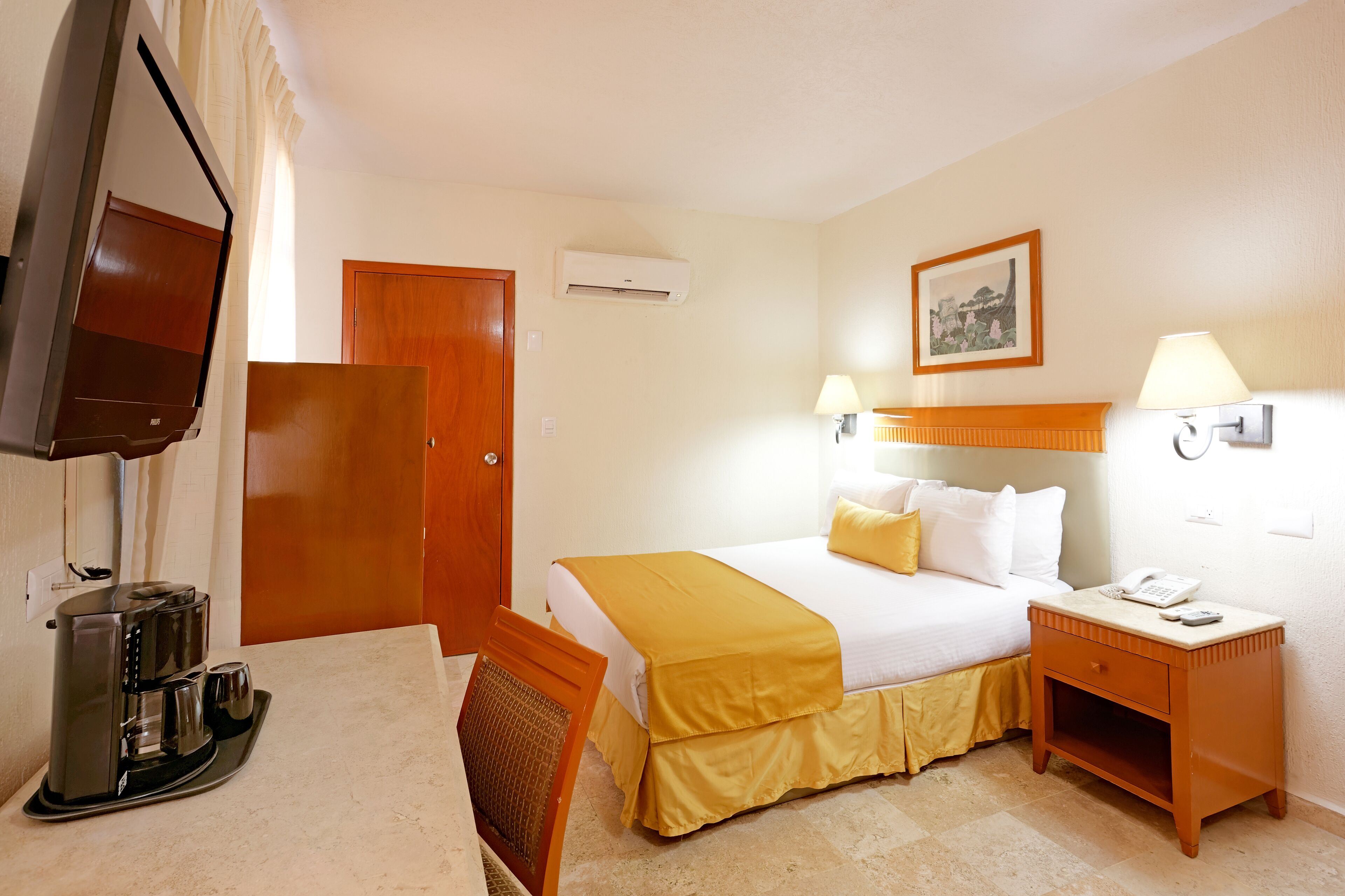 Habitación de hotel con cama, lámparas de noche, cafetera, televisor y puerta de madera.