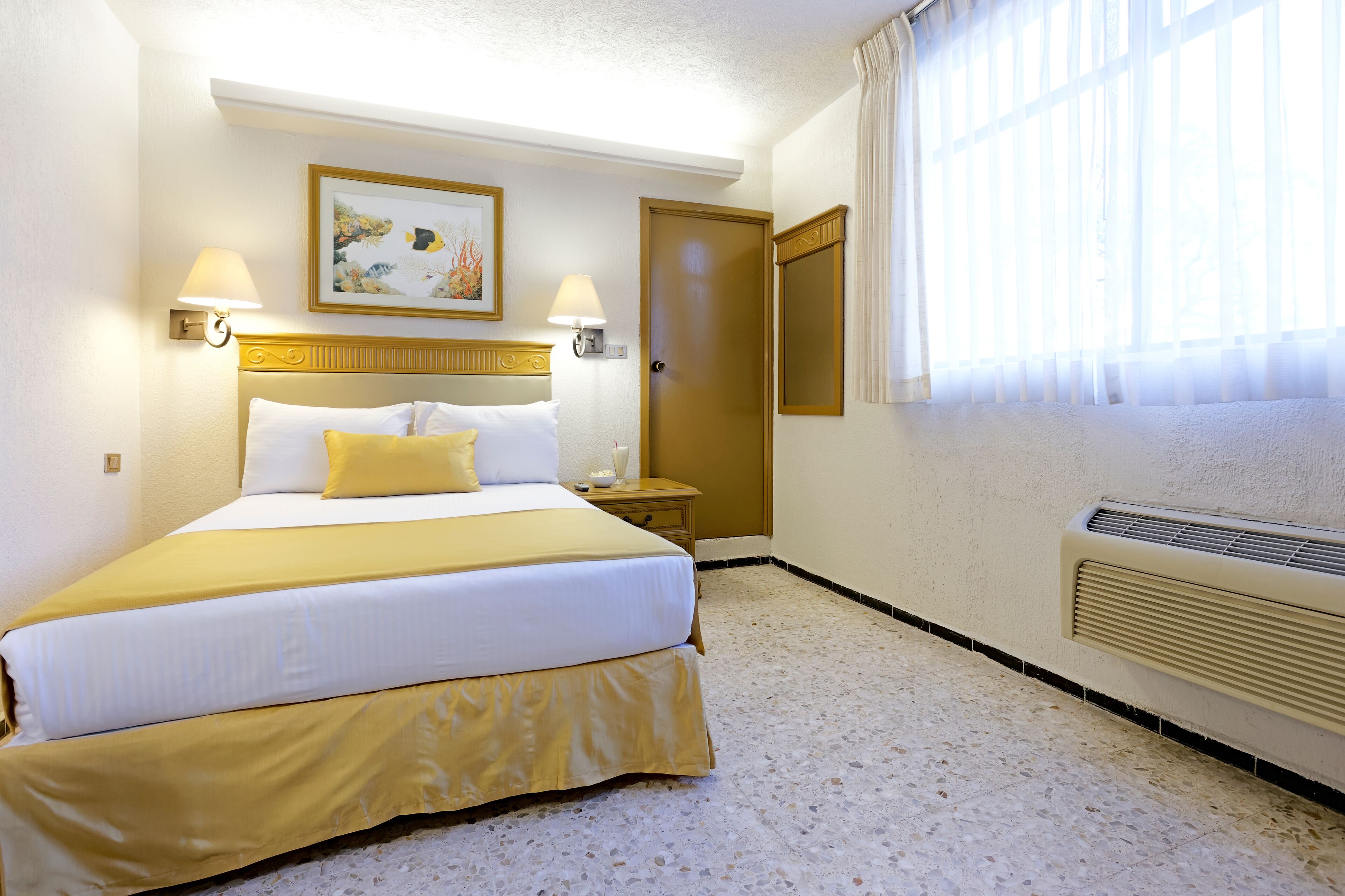A hotel room with a bed, two bedside lamps, a nightstand, a painting, a door, and a window with curtains.