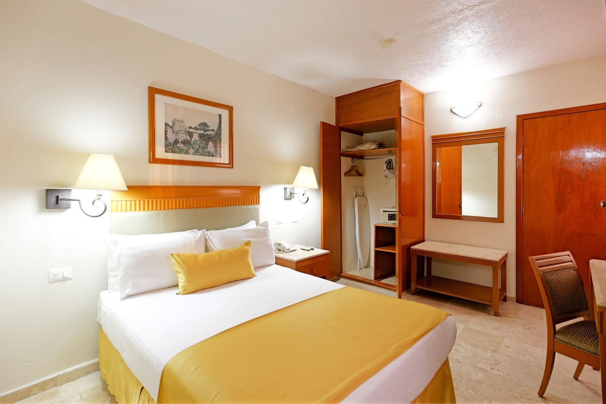 A hotel room with a bed, bedside lamps, a wooden wardrobe, a mirror, a chair, and a desk.