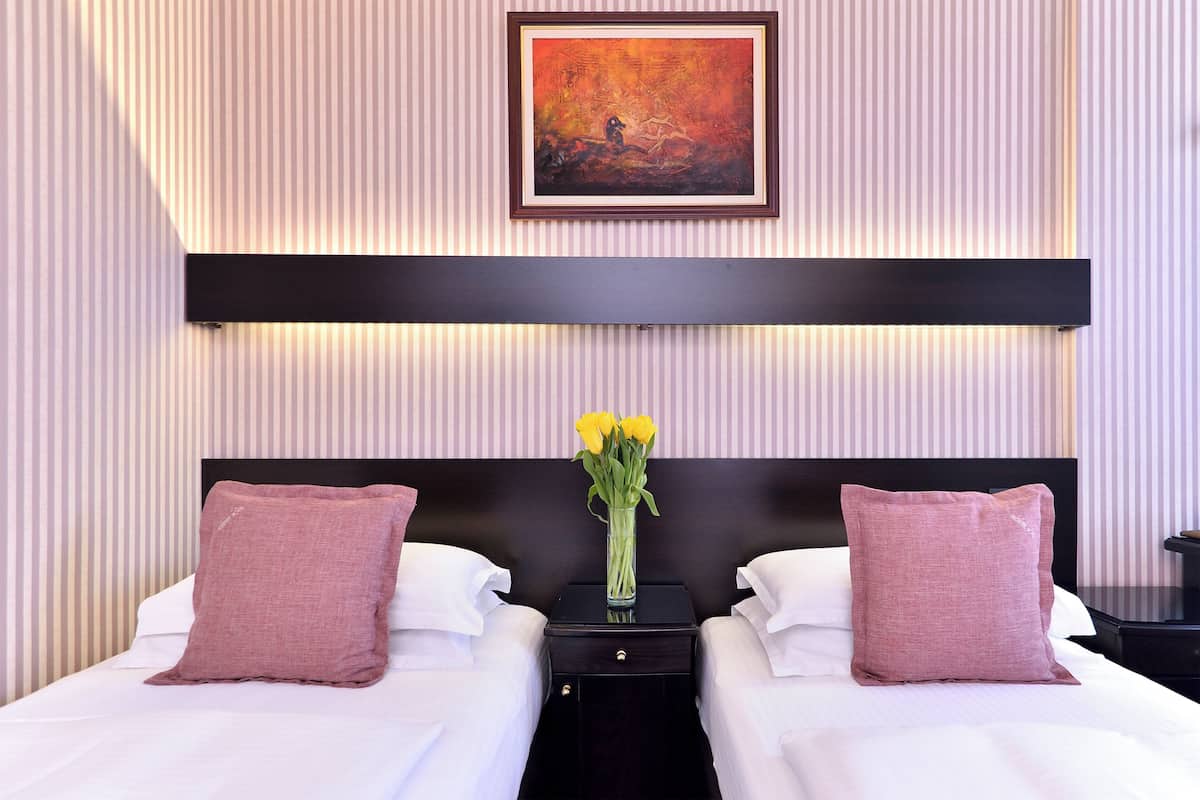 A hotel room with two beds, a nightstand with a vase of flowers, a painting on the wall, and striped wallpaper.
