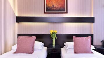 Ein Hotelzimmer mit zwei Betten, einem Nachttisch mit Blumenvase sowie einem Bild an der Wand und gestreifter Tapete.