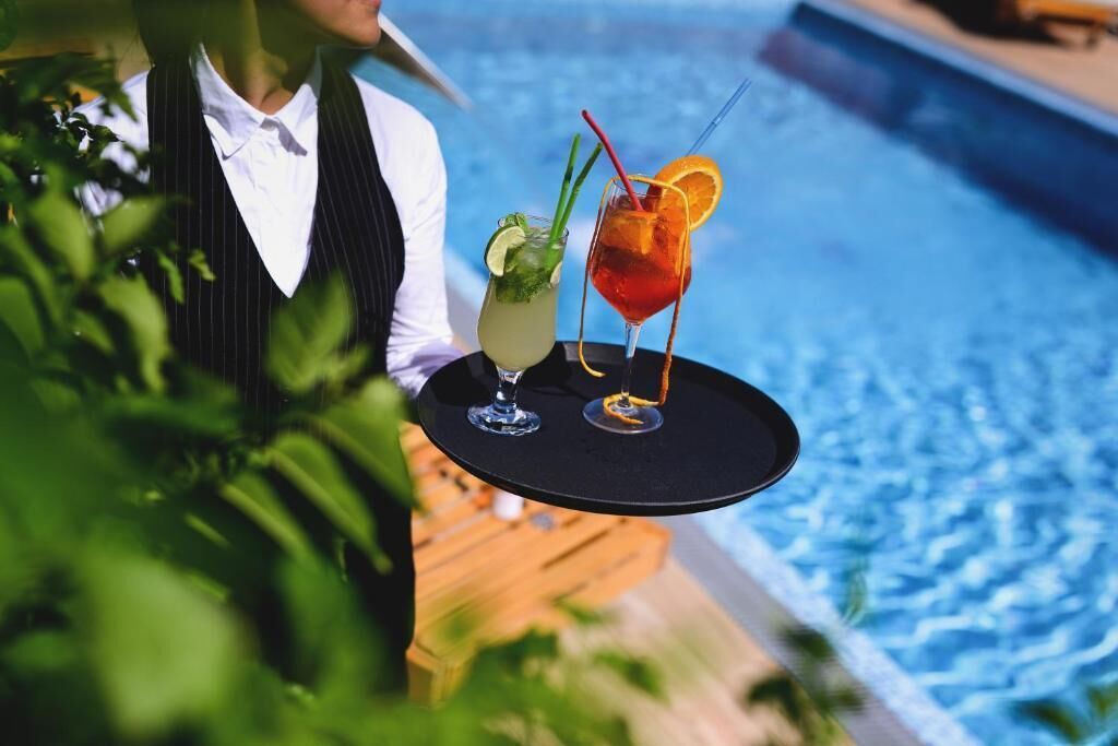 Una persona sostiene una bandeja con dos bebidas junto a una piscina.