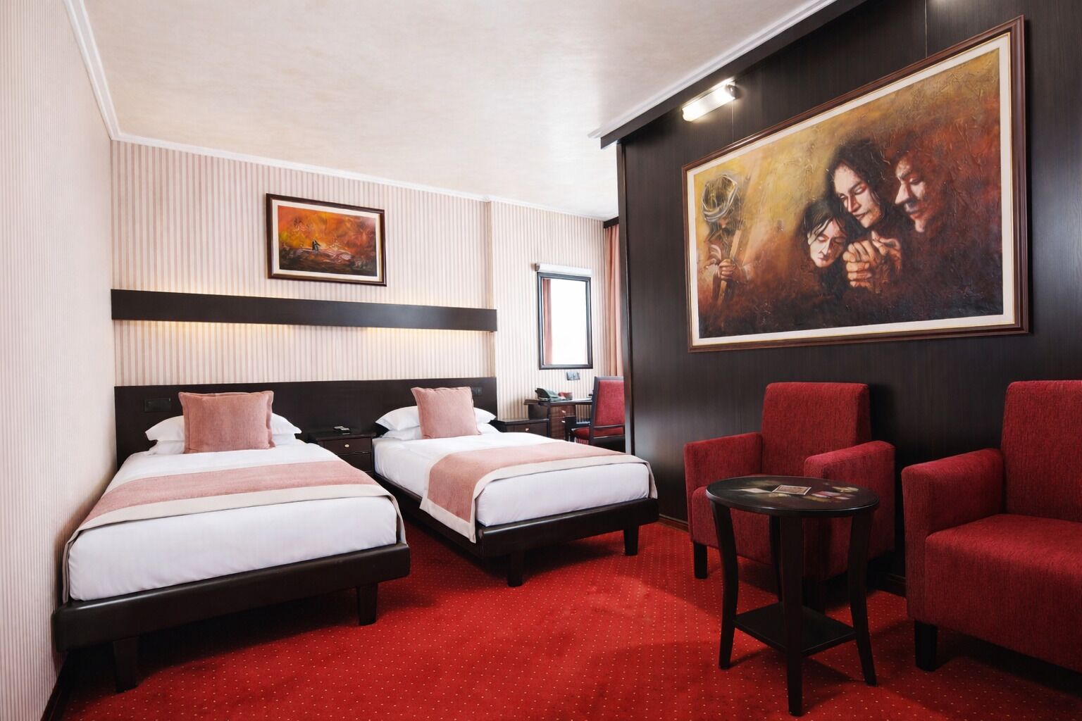 A hotel room with two beds, a red carpet, a painting, and a small table.