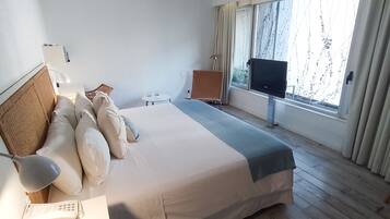 Habitación de hotel con cama, escritorio con teléfono, televisión, una mesita con vasos y ventana con cortinas.