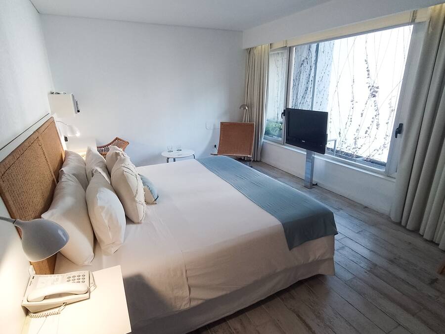 Habitación de hotel con cama, escritorio con teléfono, televisión, una mesita con vasos y ventana con cortinas.