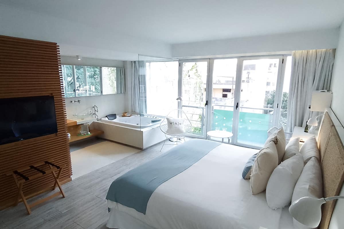 A modern hotel room with a large bed, a bathtub, a TV, and a balcony with a view.