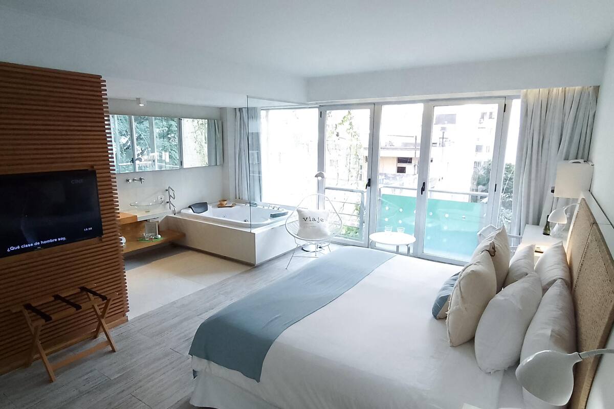 A modern hotel room with a large bed, a bathtub, a TV, and a balcony with a view.