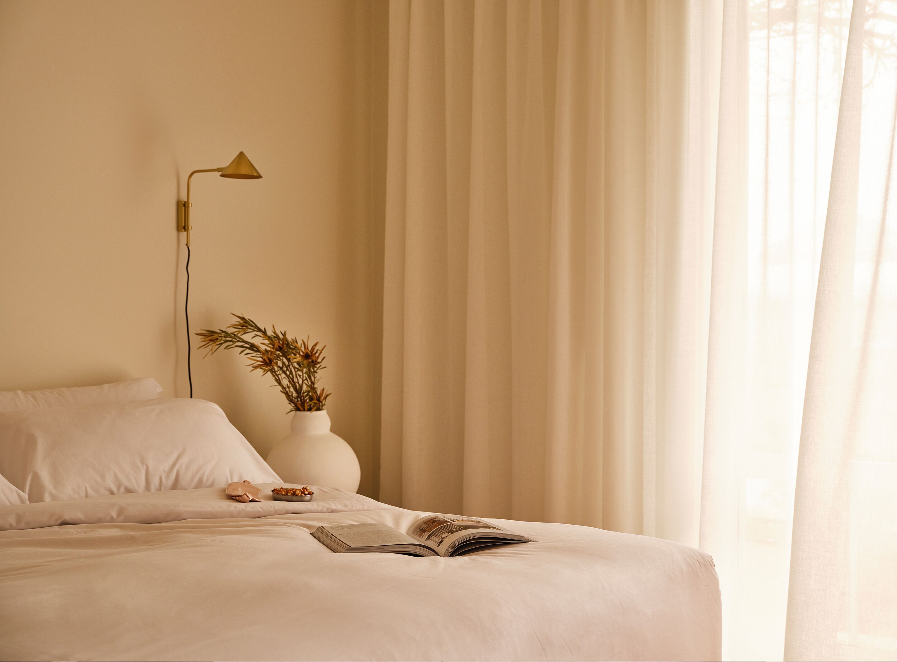 A neatly made bed with a book and a bowl of cereal, a white vase with dried plants, and a gold lamp on a bedside table.