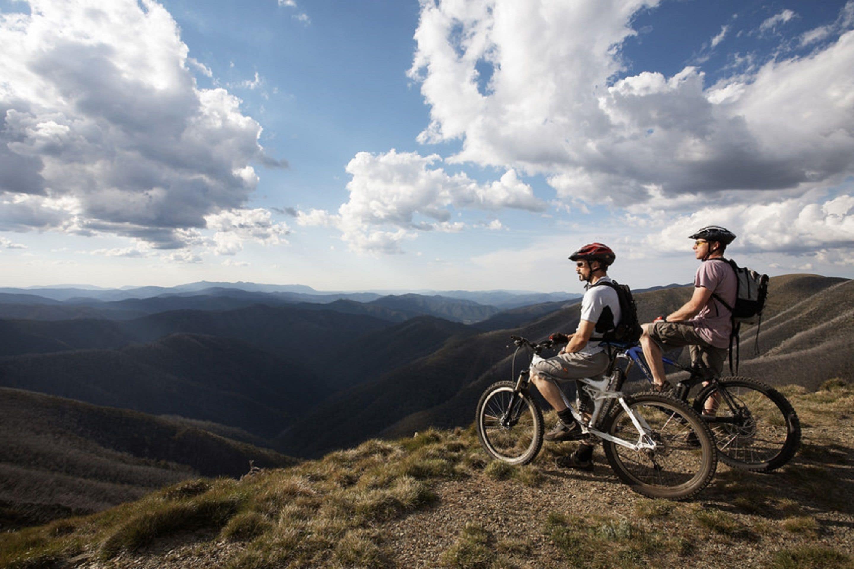 Two cyclists on a mountain trail with a view of distant mountains and a partly cloudy sky.