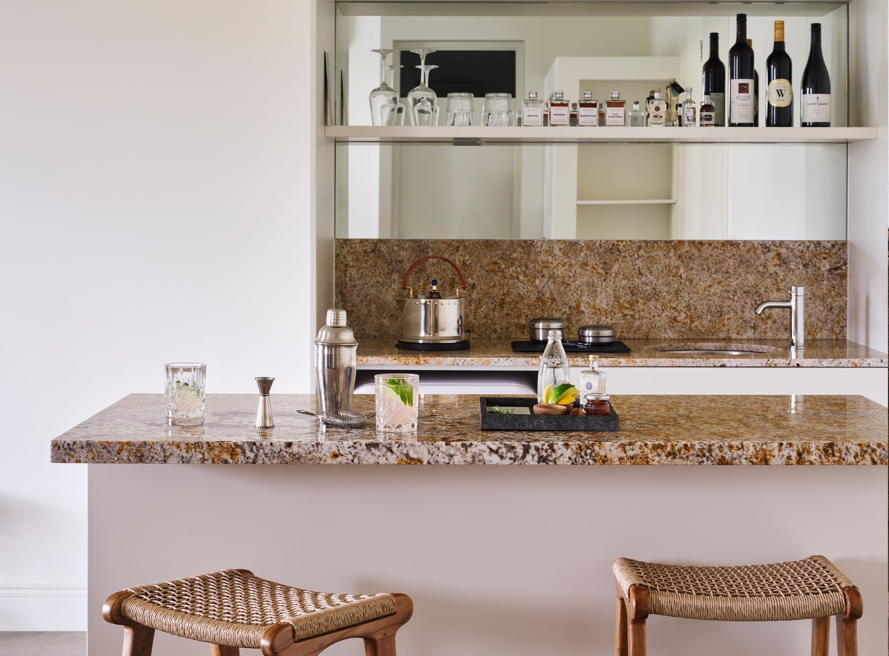 Una cocina con encimeras de granito, un fregadero de acero inoxidable y una estantería para vinos sobre la encimera. Hay dos sillas de mimbre frente a la encimera.