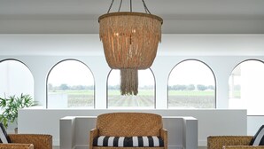 Una habitación con un sofá tejido, dos sillones, una mesa de centro de madera y una lámpara de araña grande de fibras naturales. Tres ventanas arqueadas ofrecen vistas a un campo.