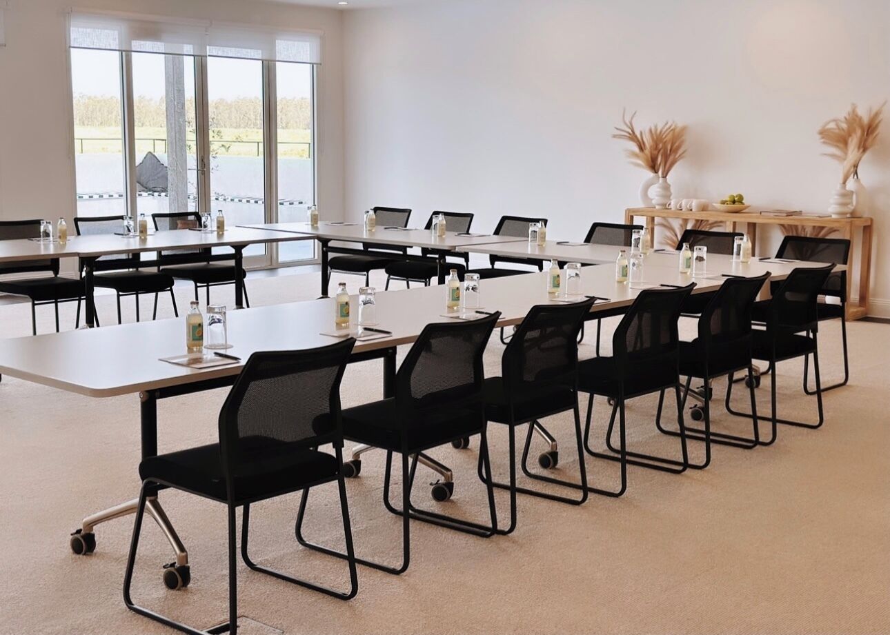 A conference room with multiple tables and chairs, large windows with a view of a parking lot, and a small decorative plant on a table.