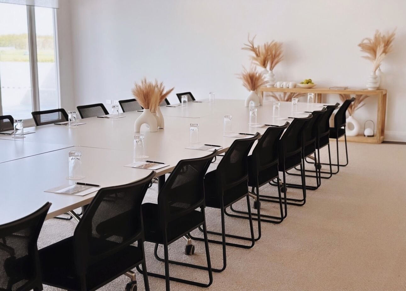 Un salón de conferencias con una larga mesa, sillas negras y jarrones blancos con plantas secas.