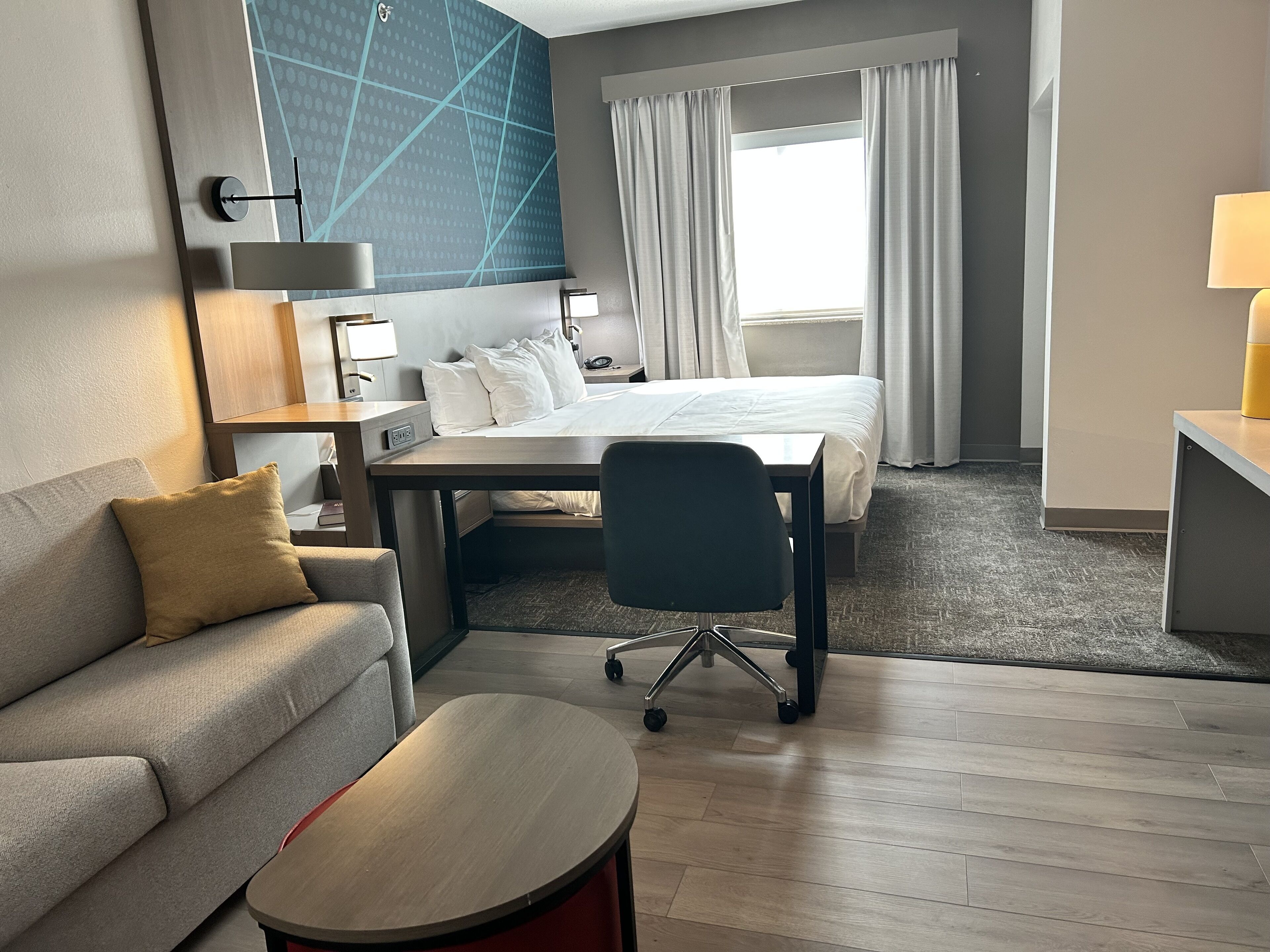 A hotel room with a bed, a desk, a chair, a sofa, and a lamp.