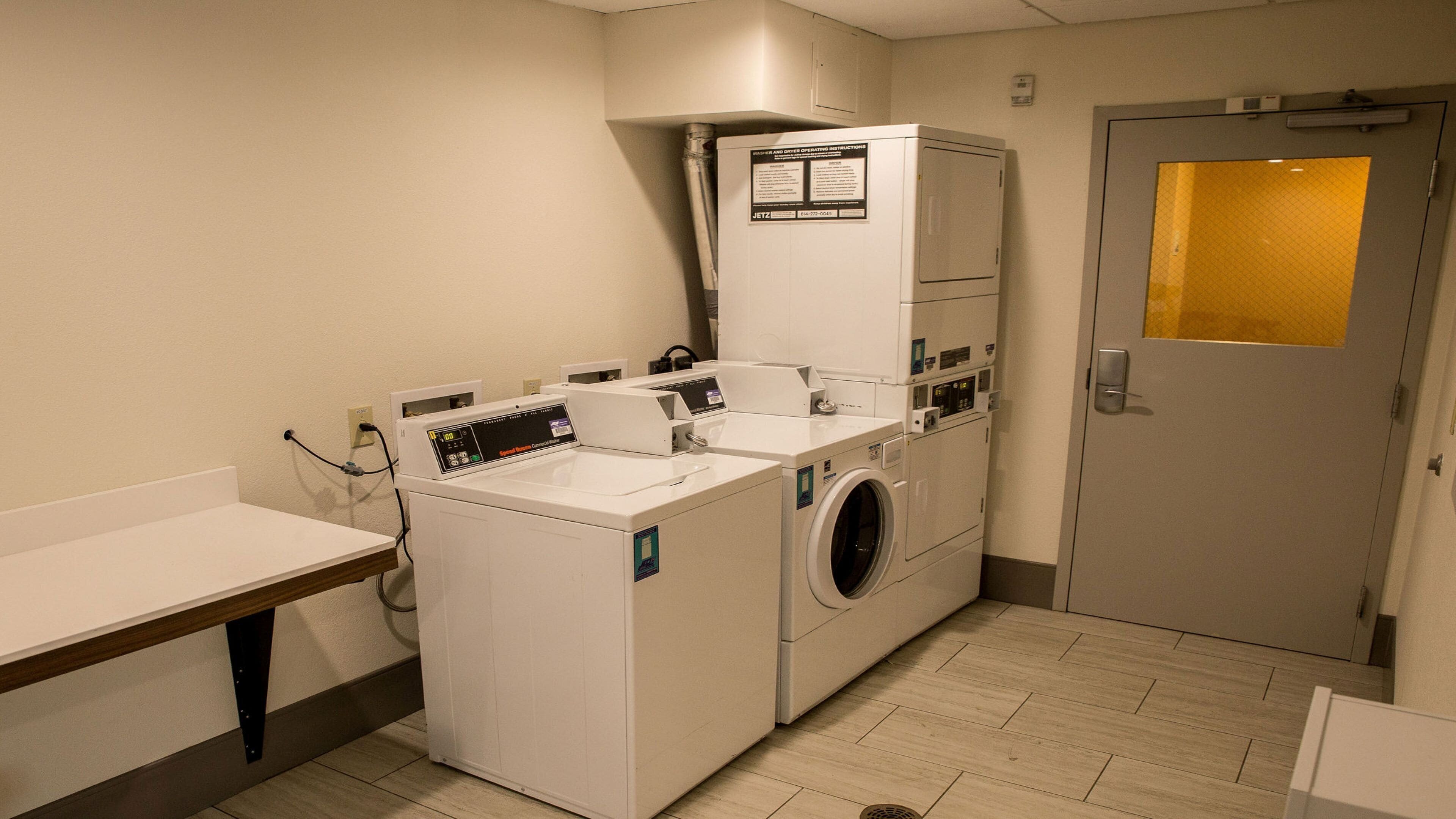 A laundry room with multiple washing machines and dryers, a white wall with a safety instruction sign, and a door with a yellow window.