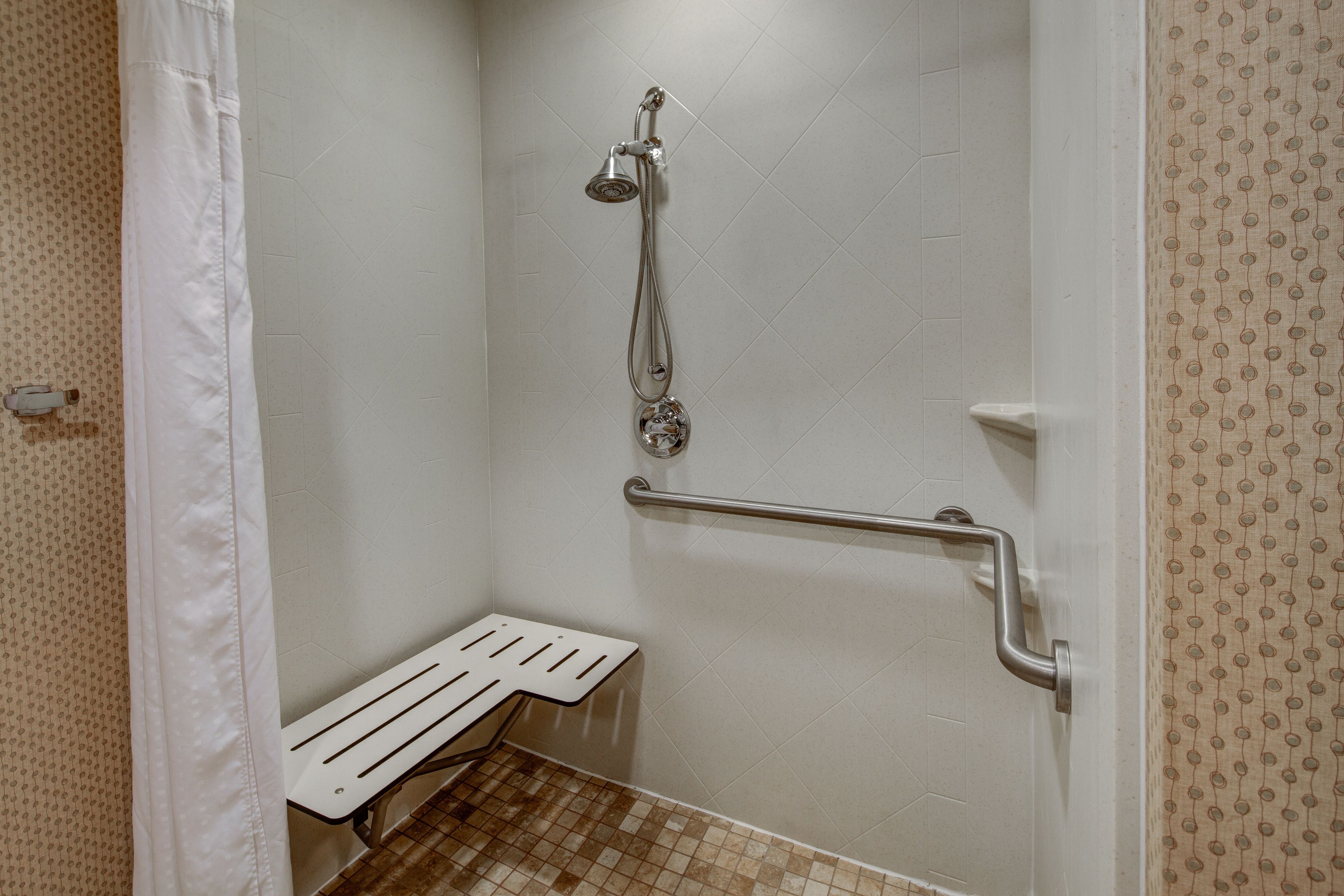 A small, white, tiled shower area with a showerhead, a grab bar, and a bench.