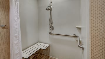 A small, white, tiled shower area with a showerhead, a grab bar, and a bench.