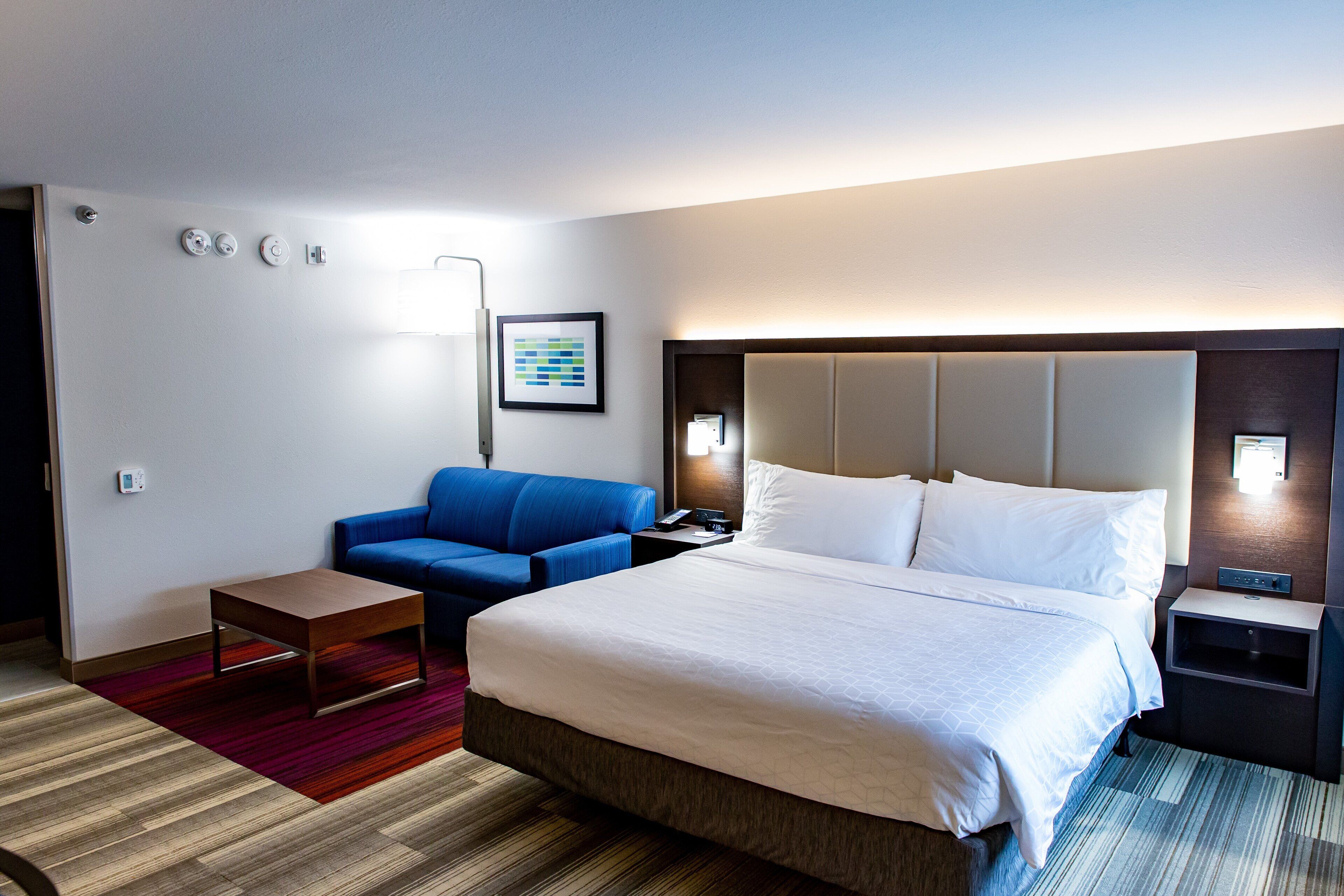 A hotel room with a large bed, a blue sofa, a small table, and a nightstand with a lamp.