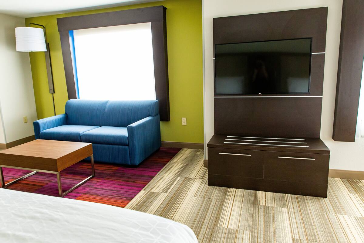 A modern hotel room with a blue sofa, a wooden coffee table, a TV mounted on a dark cabinet, and a colorful striped rug.