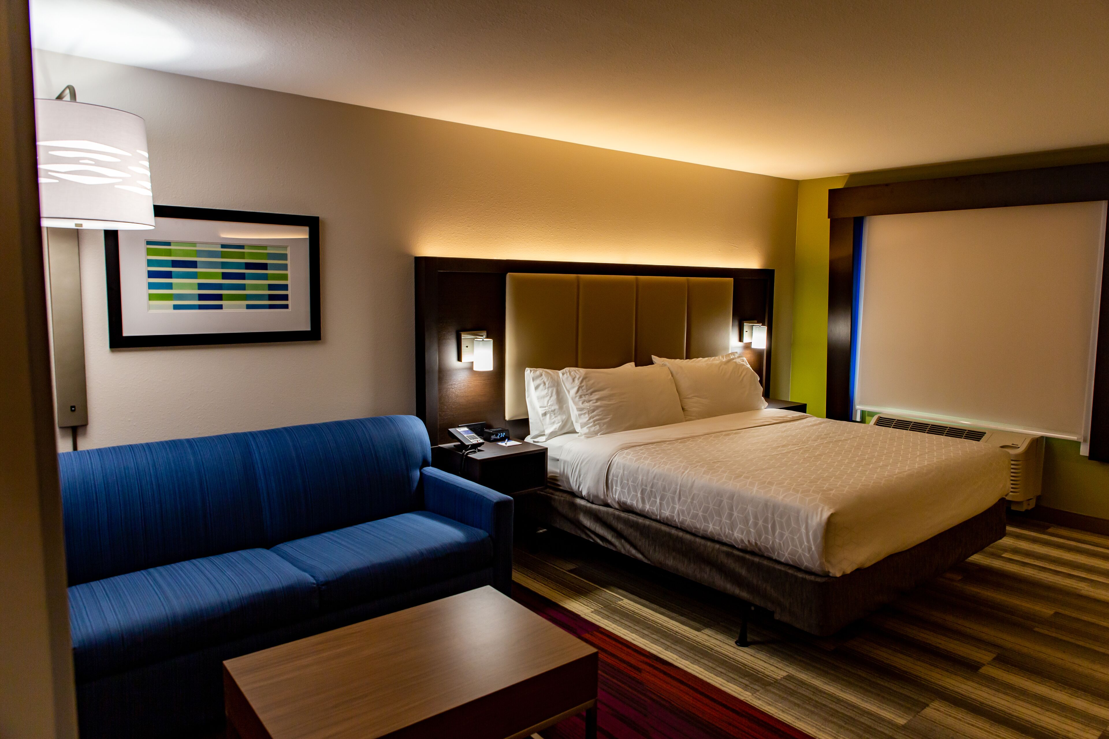 A hotel room with a bed, a blue sofa, a wooden table, a framed picture on the wall, and a window with blinds.