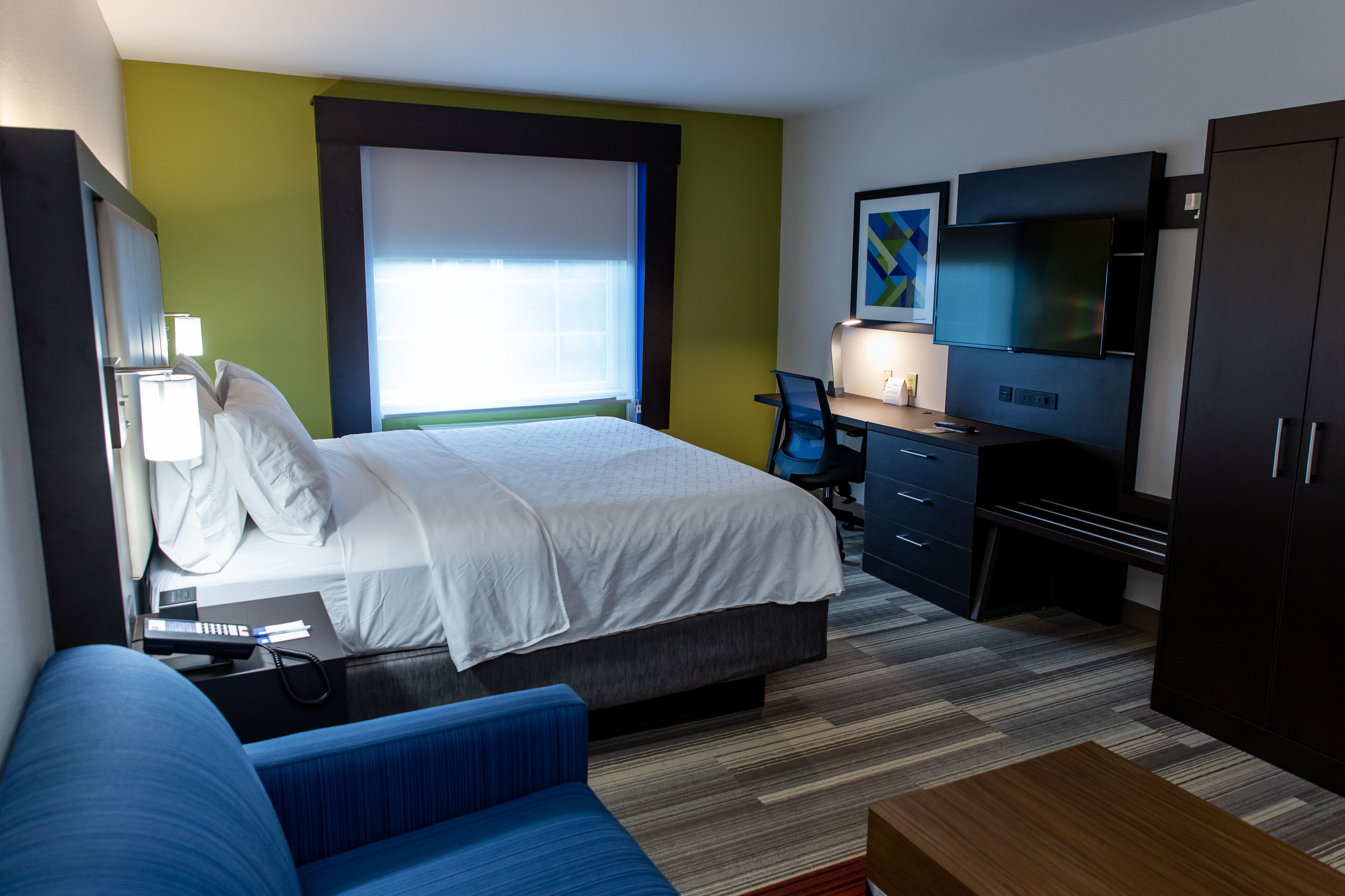 Une chambre d’hôtel équipée d’un lit, d’un bureau, d’une chaise, d’une télévision, d’une armoire et d’une fenêtre avec des stores.
