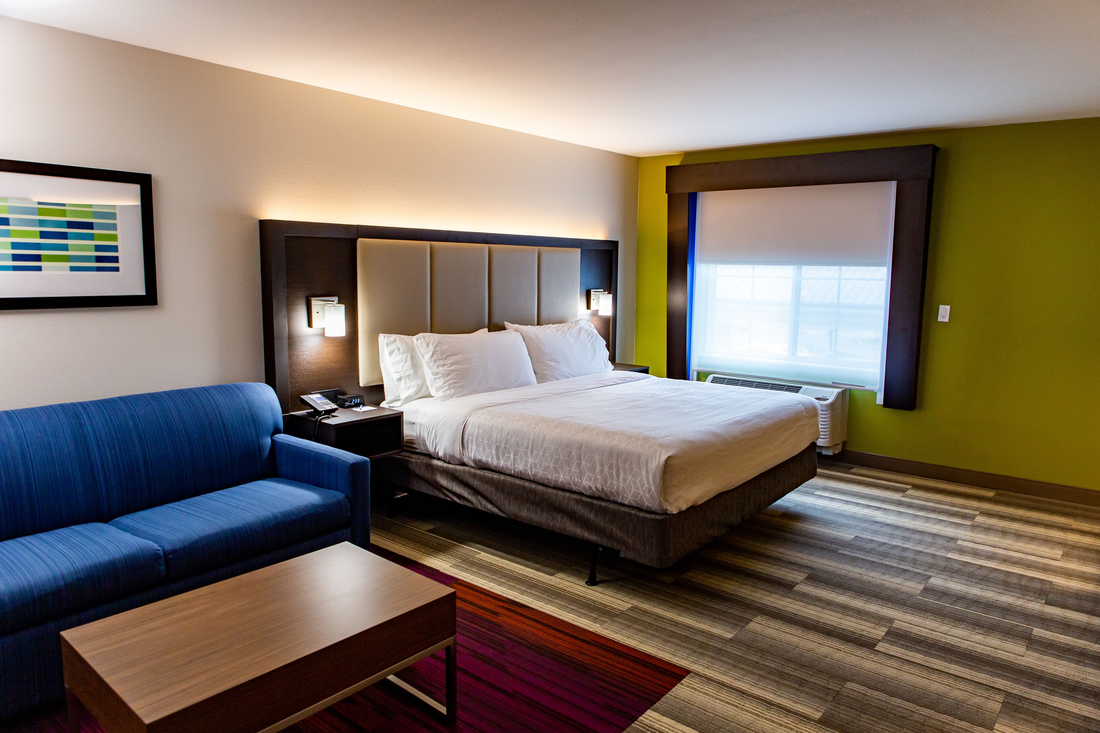 A hotel room with a bed, a blue sofa, a wooden coffee table, a nightstand, and a wall-mounted lamp.