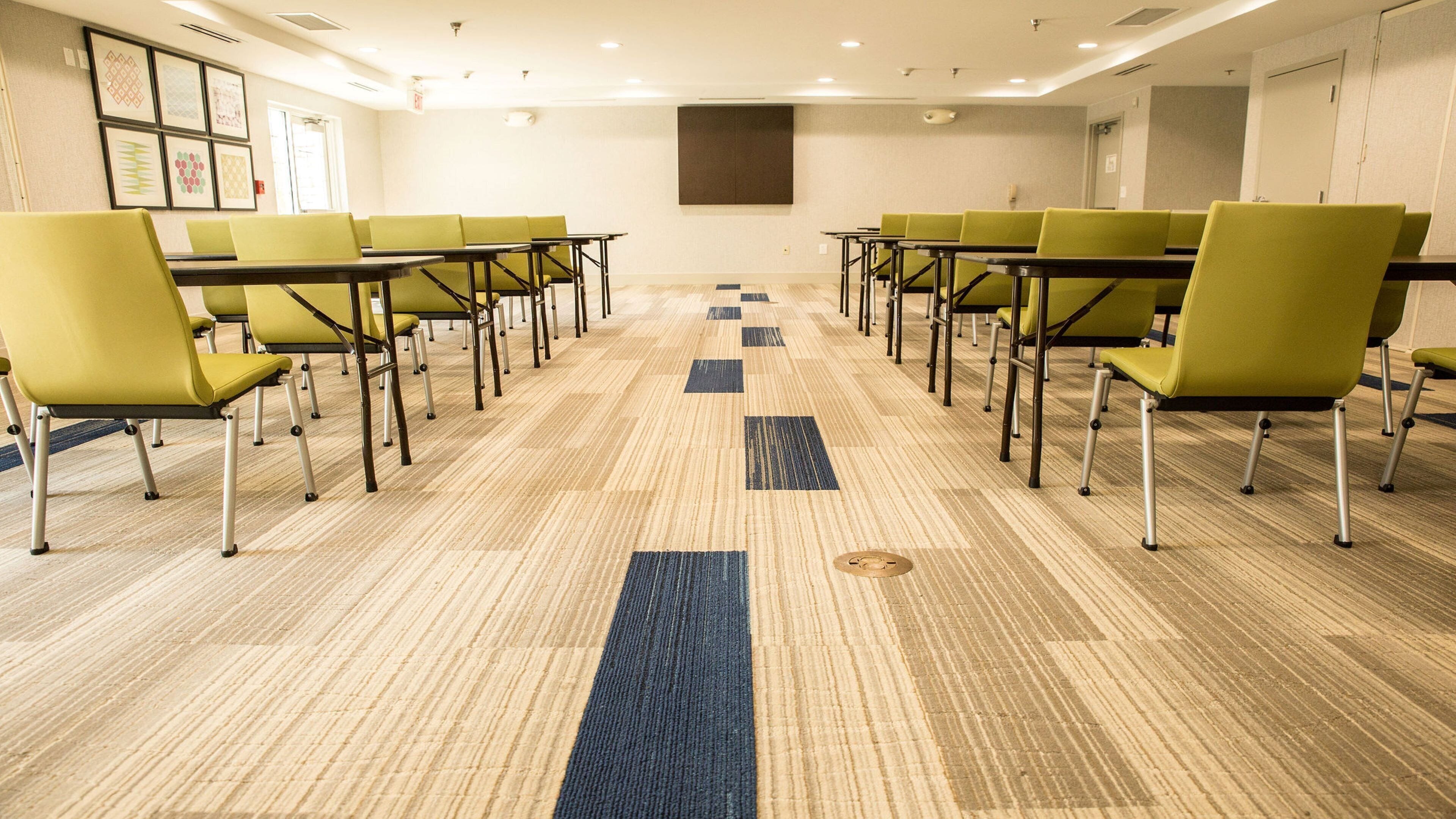 Un salón de conferencias con filas de sillas y mesas verdes, piso alfombrado y una gran pantalla instalada en la pared.