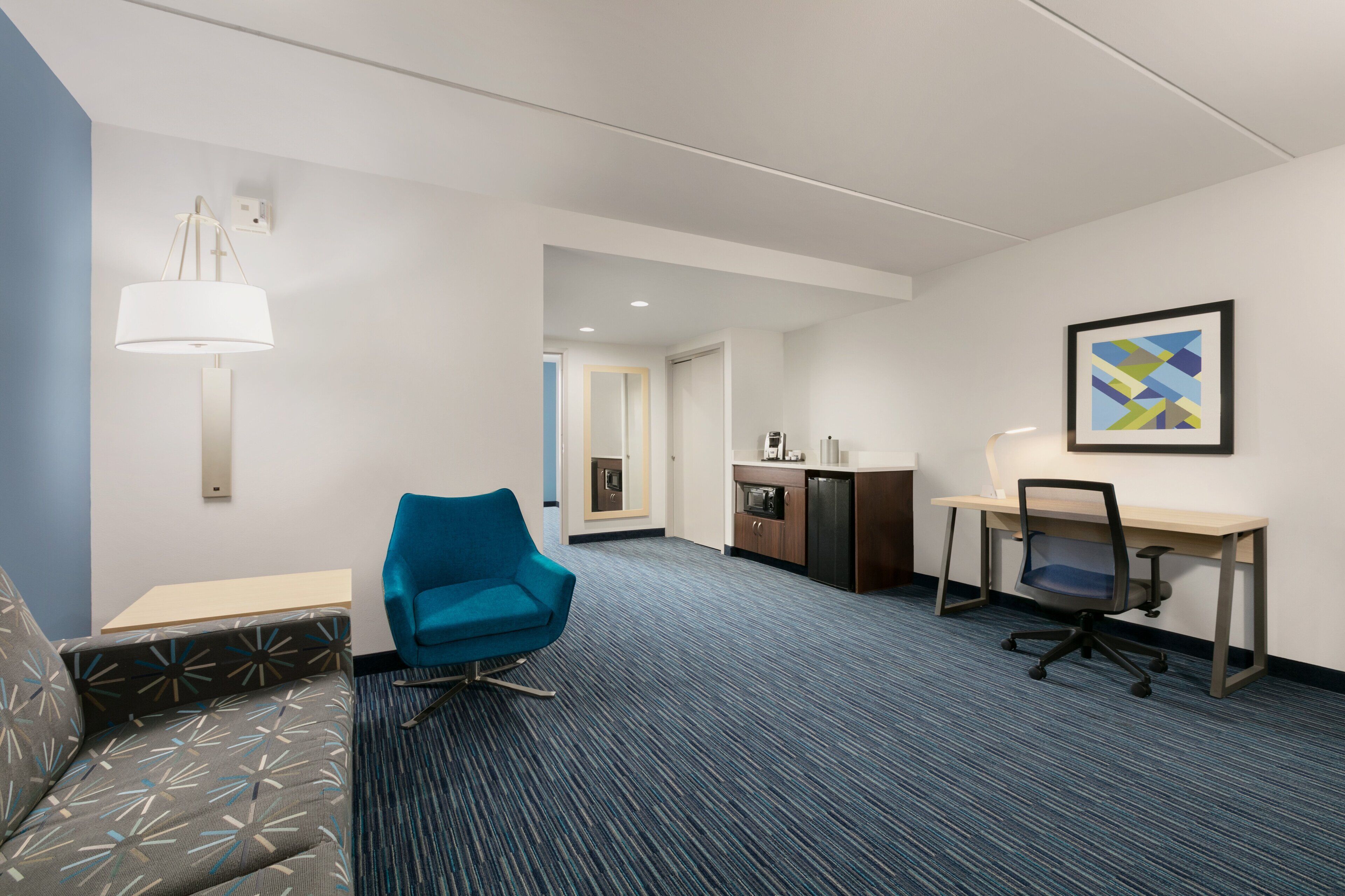 Habitación de hotel con un sillón azul, un escritorio con silla, una kitchenette y un cuadro en la pared.